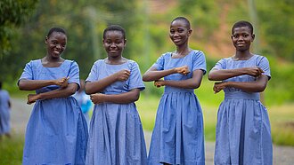 Vier ghanaische Schulmädchen in blaukarierten Schuluniformen stehen nebeneinander im Freien und lächeln in die Kamera. Sie halten ihre Unterarme vor ihrem Körper, sodass sie ein Gleichzeichen bilden.. Die Umgebung ist grün und ländlich.