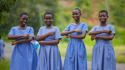 Vier ghanaische Schulmädchen in blaukarierten Schuluniformen stehen nebeneinander im Freien und lächeln in die Kamera. Sie halten ihre Unterarme vor ihrem Körper, sodass sie ein Gleichzeichen bilden.. Die Umgebung ist grün und ländlich.