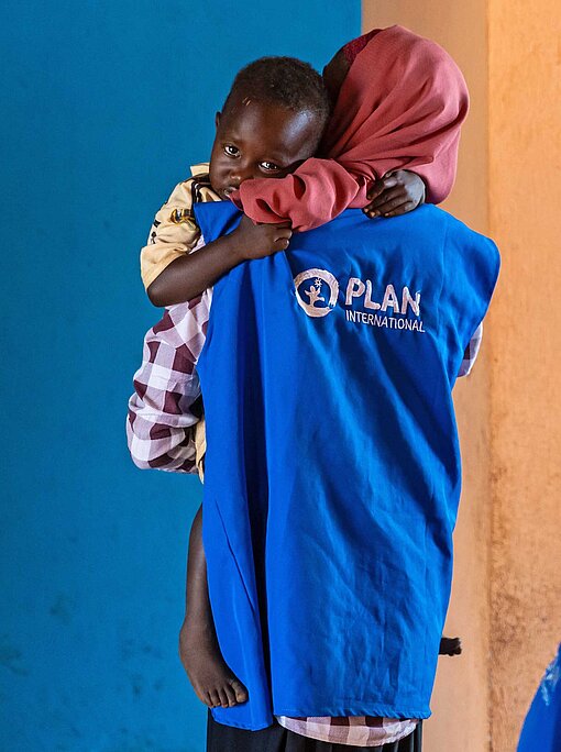 Ein Junge schmiegt sich in die Arme einer Kindergärtnerin mit dem blaueb Plan-Logo auf einer Weste