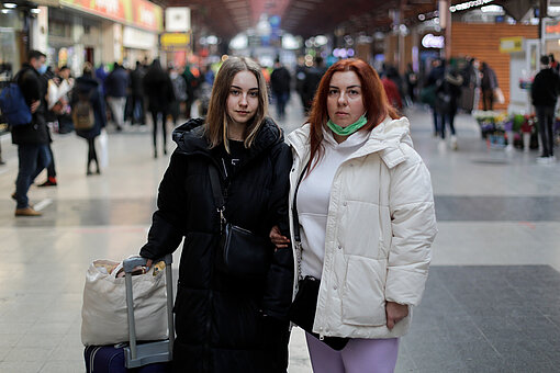 Eine Teenagerin mit Rollkoffer steht neben einer Frau auf einem Bahnhof