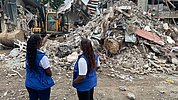 Zwei Frauen mit blauen Plan International Westen stehen vor einem großen Schutthaufen nach einem Gebäudeeinsturz. Eine Frau zeigt auf den Bagger, der im Hintergrund arbeitet.