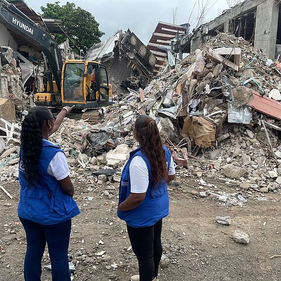 Zwei Frauen mit blauen Plan International Westen stehen vor einem großen Schutthaufen nach einem Gebäudeeinsturz. Eine Frau zeigt auf den Bagger, der im Hintergrund arbeitet.