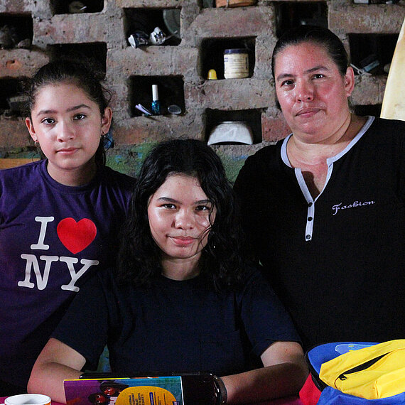 Eine Frau und zwei Mädchen, ihre Töchter, stehen an einem Tisch vor einer Backsteinwand mit kleinen Fächern, in denen verschiedene Gegenstände abgestellt sind. Auf dem Tisch befinden sich bunte Gegenstände und Taschen. Sie schauen mit einem neutralen Gesichtsausdruck in die Kamera.