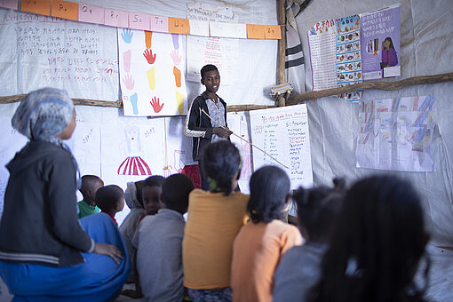 Im Jugendzentrum trat er schnell einem Life Skills Club teil. Inzwischen unterrichtet er andere Kinder. Ein Jugendlicher steht neben einem Flipchart. Vor ihm sitzen Kinder und hören ihm aufmerksam zu.