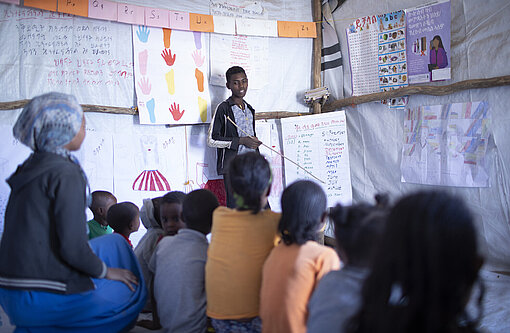 Ein Jugendlicher steht neben einem Flipchart. Vor ihm sitzen Kinder und hören ihm aufmerksam zu.