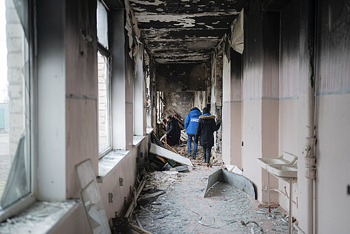 Plan International vor Ort Drei Menschen gehen durch einen durch eine Explosion zerstörten Schulflur