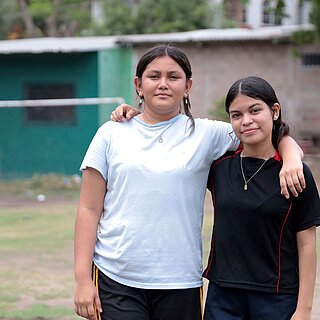 Virginia und Saidia stehen Arm in Arm draußen auf dem Fußballplatz ihrer Gemeinde