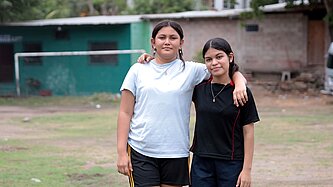 Virginia und Saidia stehen Arm in Arm draußen auf dem Fußballplatz ihrer Gemeinde