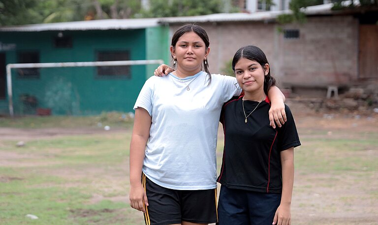 Virginia und Saidia stehen Arm in Arm draußen auf dem Fußballplatz ihrer Gemeinde