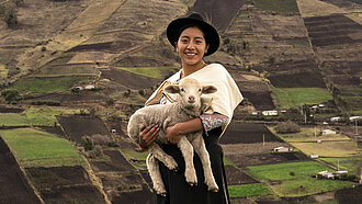 Schafe für berufliche Zukunft junger Menschen in Bolivien. Ein Mädchen steht mit einem Schaf auf dem Arm vor Feldern in Ecuador.
