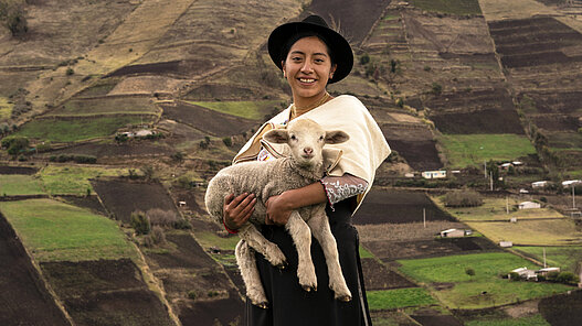 Ein Mädchen steht mit einem Schaf auf dem Arm vor Feldern in Ecuador.