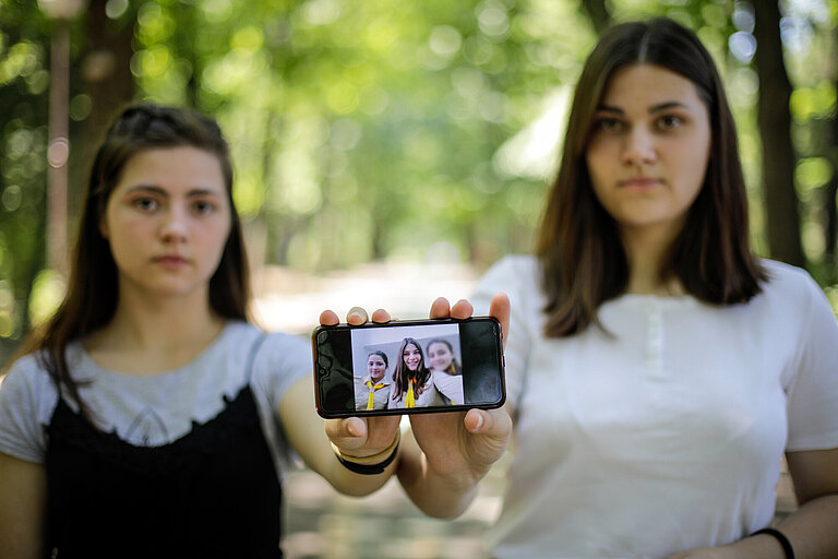 Zwei junge Frauen halten ein Handy in die Kamera, auf dem Display sieht man ein Foto mit mehreren Personen darauf