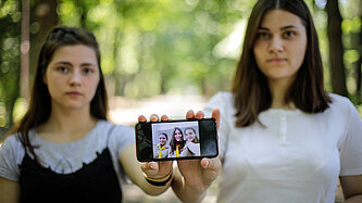 Zwei junge Frauen stehen nebeneinander und zeigen ein Handy in die Kamera, auf dem ein Bild mit drei Menschen zu sehen ist