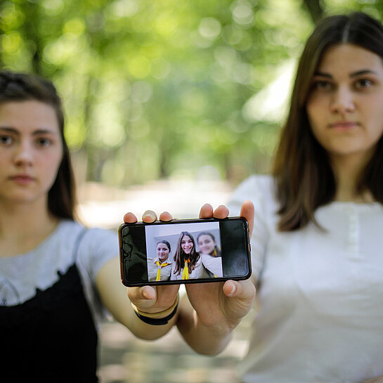 Zwei junge Frauen stehen nebeneinander und zeigen ein Handy in die Kamera, auf dem ein Bild mit drei Menschen zu sehen ist