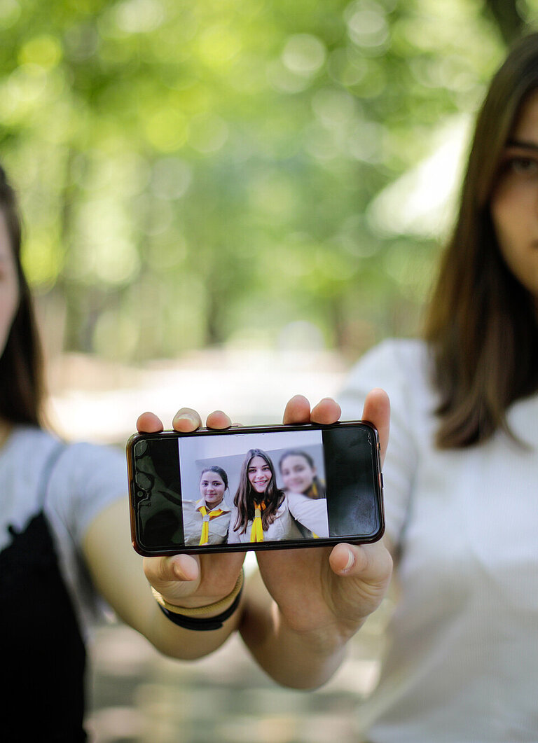 Zwei junge Frauen stehen nebeneinander und zeigen ein Handy in die Kamera, auf dem ein Bild mit drei Menschen zu sehen ist