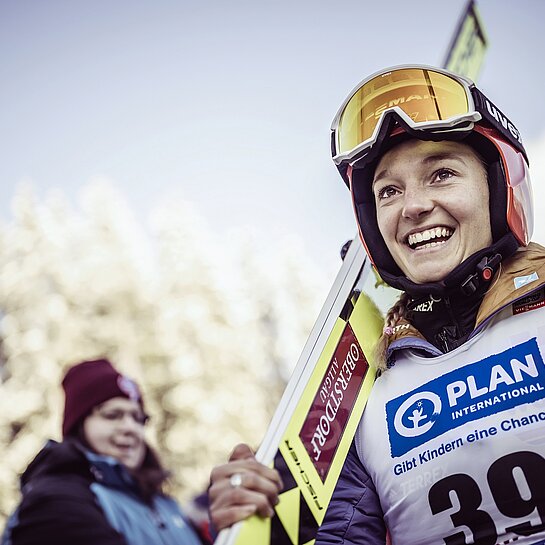 Eine Skispringerin in Wettkampfkleidung trägt ihre Ski auf der rechten Schulter. Sie lacht.