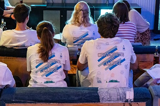 Mehrere junge Menschen sitzen in Reihen und schauen nach vorn, man sieht sie von hinten, auf ihren Shirt steht Champions of Change