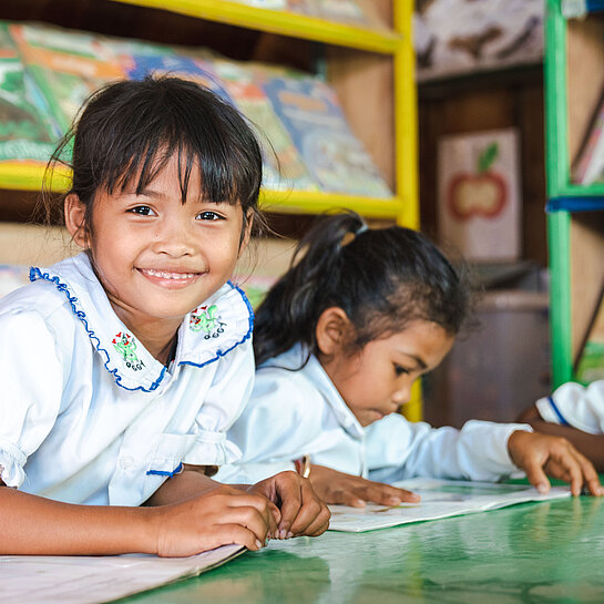 Lächelndes Mädchen liest in einem Kinderbuch, umgeben von weiteren Kindern in einer Lernumgebung in Kambodscha