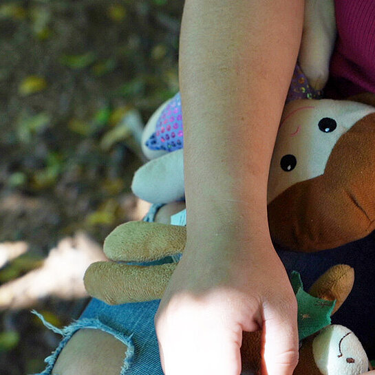 Nahaufnahme einer Person, die ihre Hand auf ihrem Bein abgelegt hat und unter dem Unterarm klemmt eine Stoffpuppe