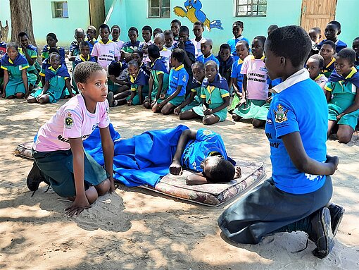 Theaterstück als Malaria-Aufklärung Vier Schüler:innen führen vor ihren Mitschüler:innen ein Theaterstück auf, zwei von ihnen liegen auf einer Matratze, die anderen beiden knien daneben