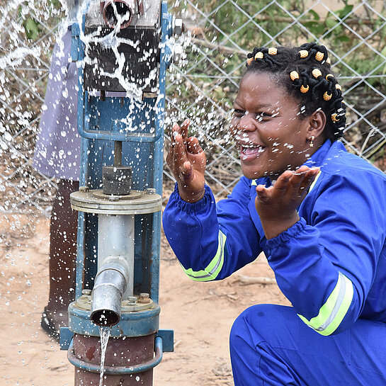 Nomsa, eine junge Frau, hockt vor einer Wasserpumpe und spritzt spielerisch mit Wasser.