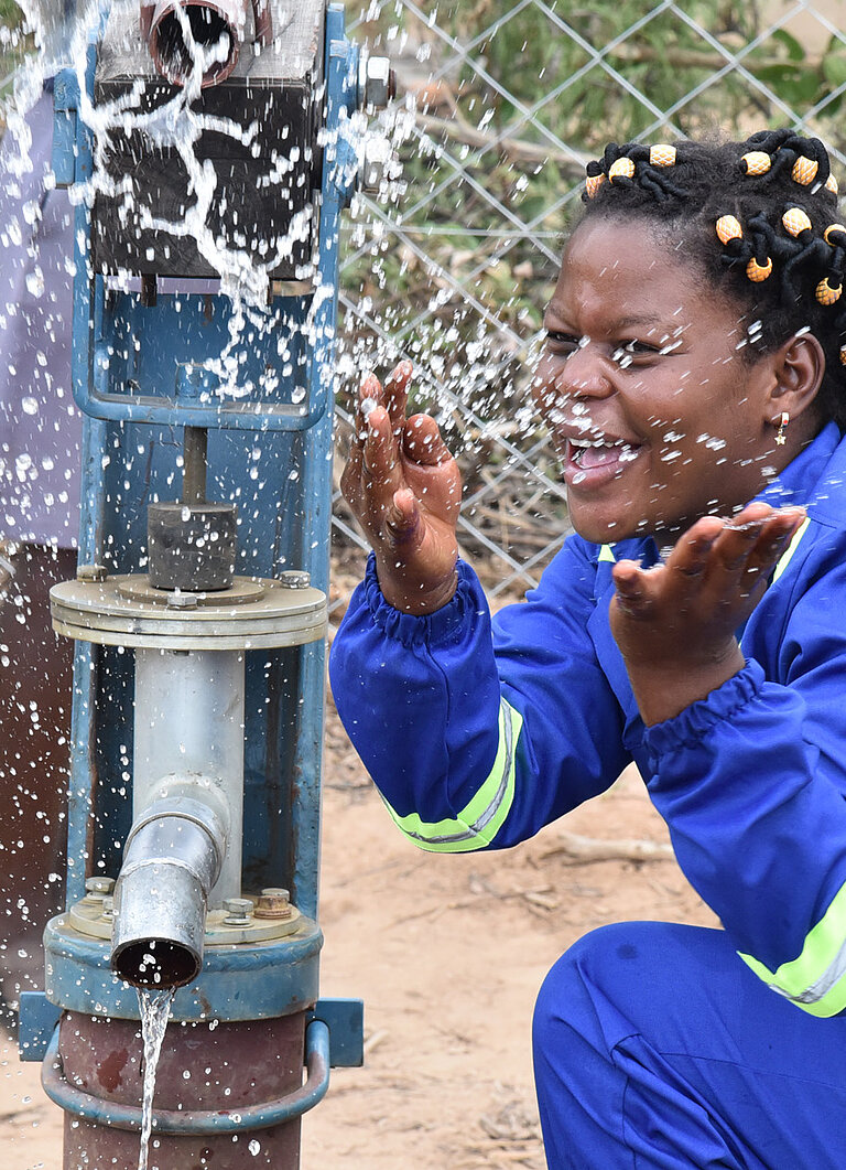 Nomsa, eine junge Frau, hockt vor einer Wasserpumpe und spritzt spielerisch mit Wasser.
