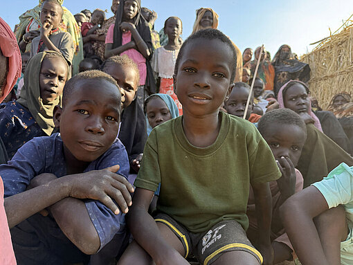 Mehrere Kinder stehen und sitzen in einem Camp in Tawila. Einige schauen in die Kamera, im Hintergrund sind Notunterkünfte aus Hirsegras zu sehen.