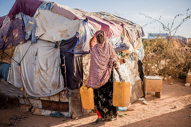 Eine Frau hält zwei große Wasserkanister in den Händen. Sie steht vor einem Zelt.
