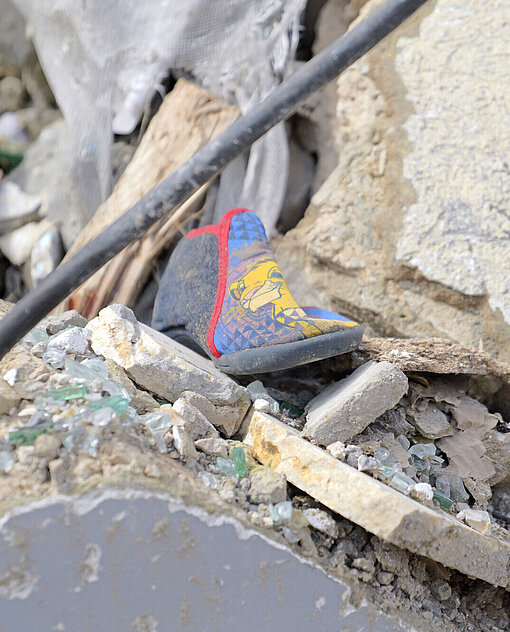 Nahaufnahme von Trümmern mit einem Kinderschuh, der zwischen Scherben und zerbrochenem Material liegt. Kabel und Baufragmente sind verstreut sichtbar.