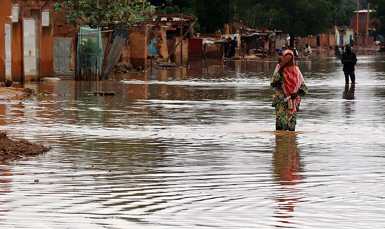 Eine junge Frau in buntem Gewand läuft durch eine überflutete Straße. Sie hält ihre Schuhe in der Hand.