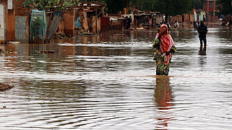Eine junge Frau in buntem Gewand läuft durch eine überflutete Straße. Sie hält ihre Schuhe in der Hand.