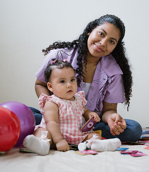 Eine junge Frau sitzt mit einem Kleinkind auf dem Boden, umgeben von bunten Ballons und Spielkarten. Beide wirken konzentriert auf die Spielsachen.