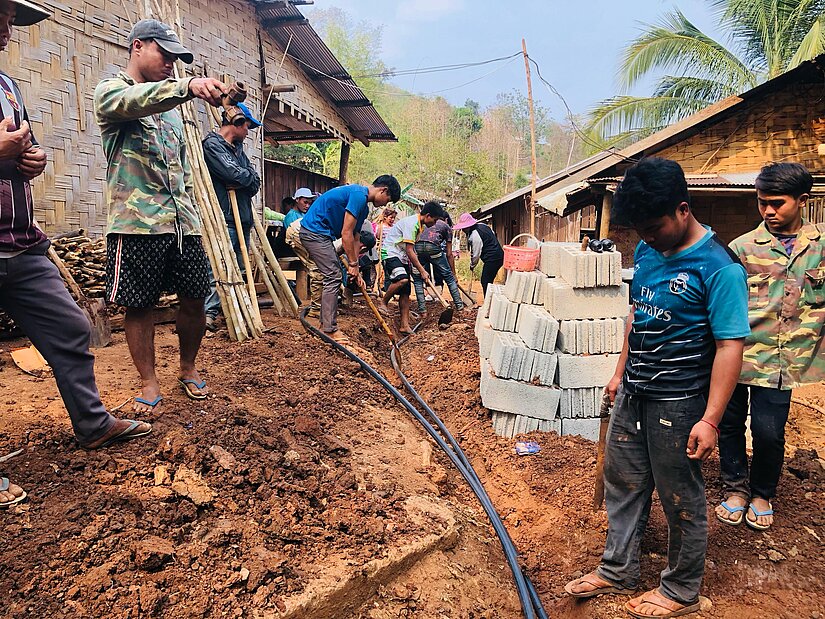 Mehrere Männer und Jungen arbeiten gemeinsam an einer Baustelle in einem Dorf, umgeben von Bambushäusern und Baumaterial wie Ziegelsteinen. Sie verlegen Rohre im aufgegrabenen Boden; einige halten Werkzeuge, andere beobachten. 