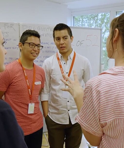 Zwei junge Männer stehen vor einer Tafel und sprechen mit einer jungen Frau.