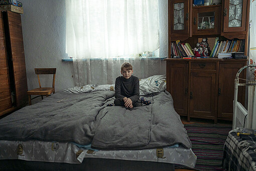 Ivan (11) sitzt auf einem Bett in dem Schlafzimmer, das er sich mit seinen Geschwistern teilt. Ein junge sitzt im Schneidersitz ein wenig verloren mitten auf einem Bett.