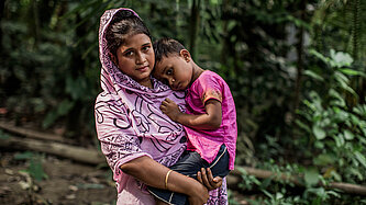 Eine junge Frau steht draußen im Grünen. Auf dem Arm trägt sie ein Kleinkind, das sich an sie lehnt. Beide schauen neutral bis traurig.