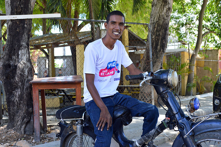 Carlos sitzt auf seinem Motorrad und lächelt in die Kamera. Er trägt ein weißes T-Shirt, das mit einem Schriftzug bedruckt ist.