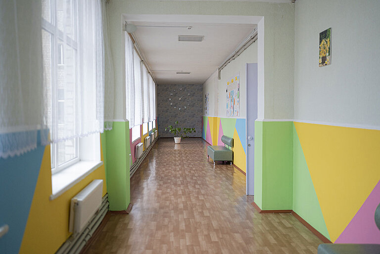 Ein leerer, lichtdurchfluteter Korridor mit Holzboden, dessen Wände in hellen, bunten Farben gestrichen sind. 