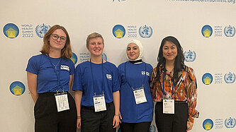 Vier junge Menschen stehen in Plan T-Shirts vor der Pressewand der Weltgesundheitskonferenz