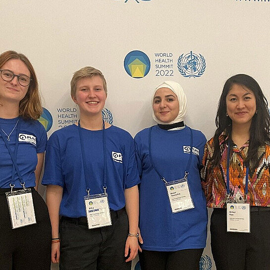 Vier junge Menschen stehen in Plan T-Shirts vor der Pressewand der Weltgesundheitskonferenz