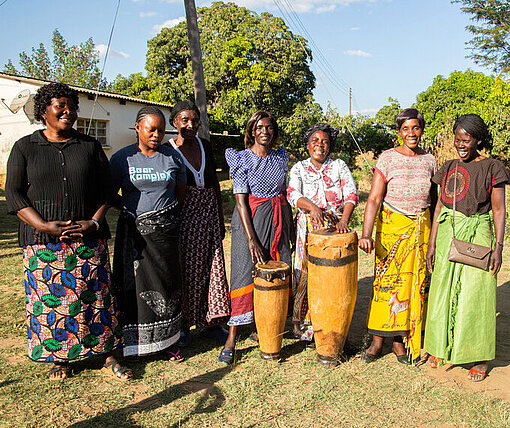 Eine Gruppe Frauen und ein Mann stehen im Halbkreis auf einer Wiese; zwei Trommeln sind in ihrer Mitte aufgestellt