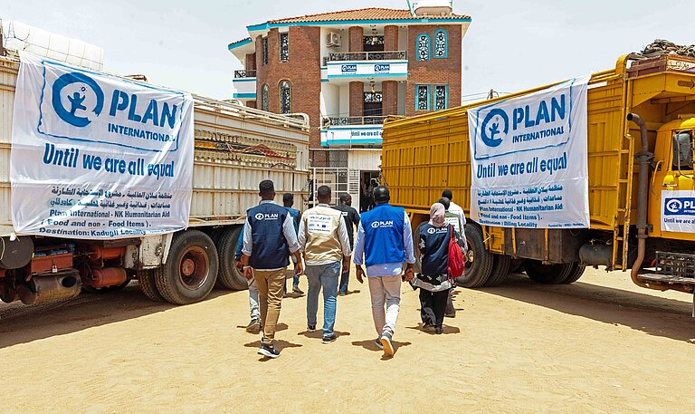 Personen mit Plan-Logo auf der Bekleidung laufen zwischen zwei LKW mit humanitärer Hilfe hindurch.