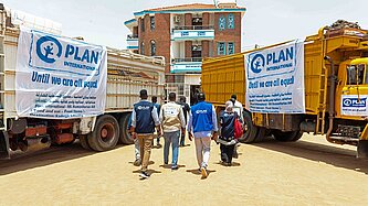 Personen mit Plan-Logo auf der Bekleidung laufen zwischen zwei LKW mit humanitärer Hilfe hindurch.