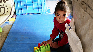 Ein Mädchen aus Syrien sitzt in einem Geflüchtetencamp auf dem Boden und spielt mit Spielzeug. Sie schaut direkt in die Kamera.