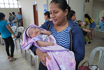 Eine junge Frau hält ein Baby im Arm, beide in hellen Farben gekleidet. Die junge Frau trägt eine blau gemusterte Bluse und schaut fürsorglich und konzentriert auf das Kind. Im Hintergrund sind weitere Erwachsene, Jugendliche und kleine Kinder zu sehen, die miteinander sprechen oder spielen.