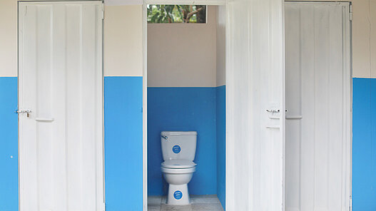 Die Tür einer Toilettenkabine steht offen. Links und rechts davon befinden sich weitere geschlossene Kabinen. 