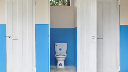 Die Tür einer Toilettenkabine steht offen. Links und rechts davon befinden sich weitere geschlossene Kabinen. 