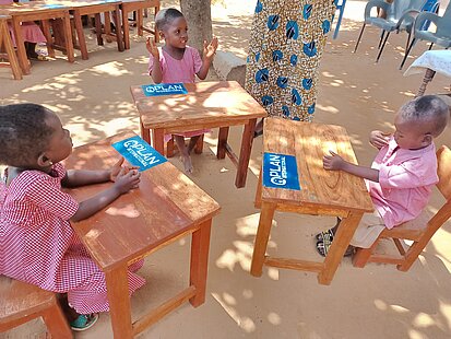 Kleinkinder sitzen an Schultischen mit Plan-Logos