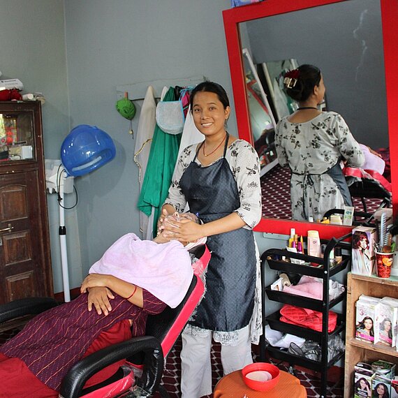 Eine junge Frau steht in einem Friseursalon und behandelt eine Kundin, die in einem Friseurstuhl sitzt. Im Hintergrund sind Friseurausrüstung und Produkte sichtbar.