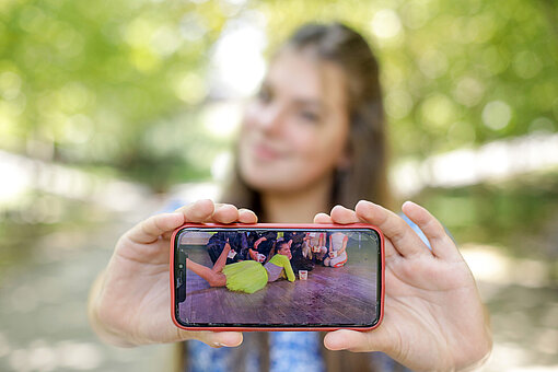 Ein Mädchen hält ein Handy in die Kamera, auf dem Display sieht man ein Foto von ihr beim Tanzen.